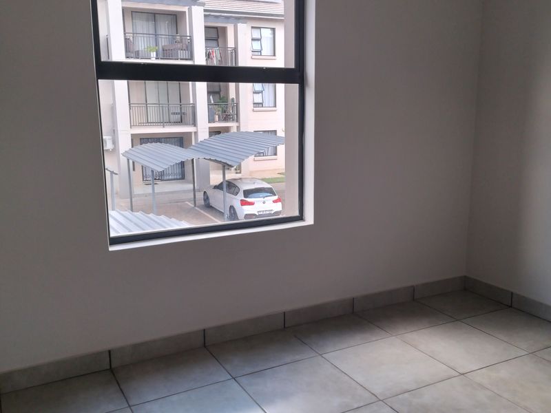 A bright, empty bedroom featuring neutral tiled flooring and a large window overlooking the apartment complex parking area.