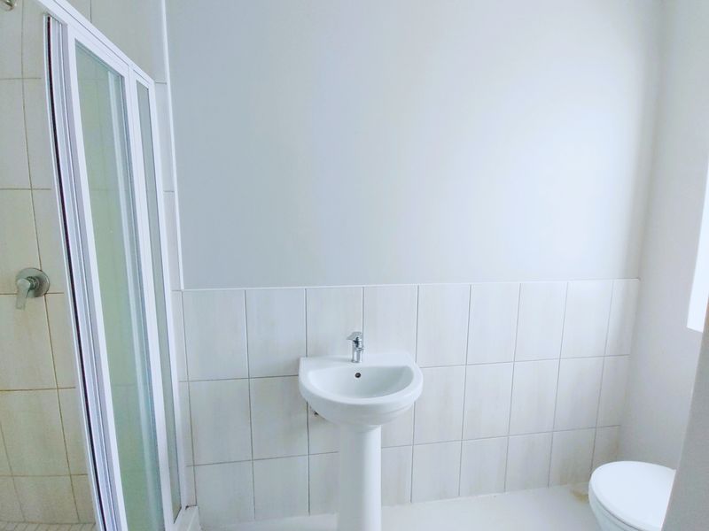 A clean, minimalist bathroom featuring a pedestal basin, a shower enclosure, and a toilet with neutral wall tiling.