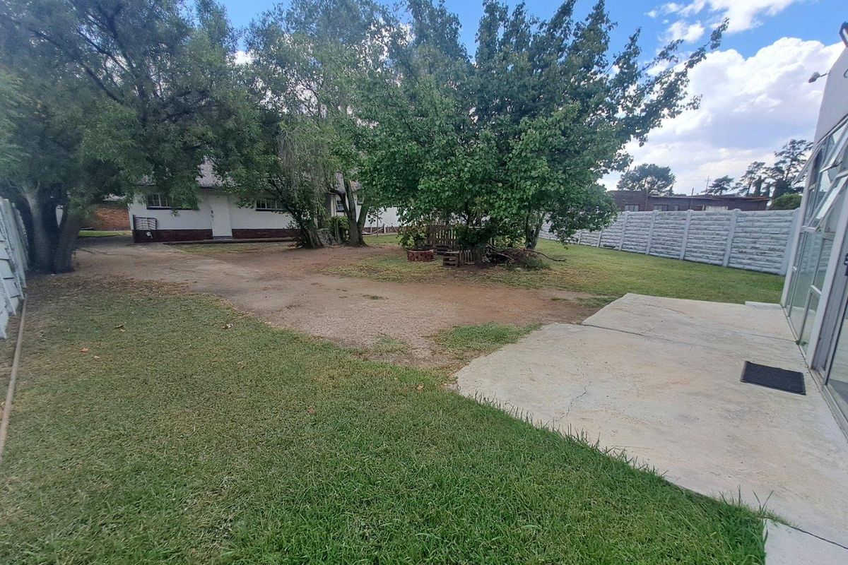A spacious rear garden featuring a concrete patio area, mature trees, and perimeter precast concrete walling.