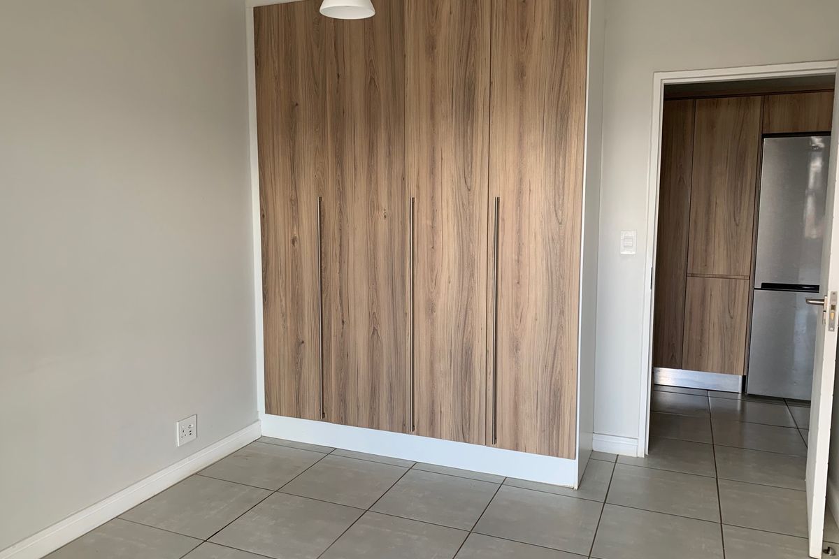 A clean, empty bedroom featuring modern wood-finish built-in cupboards and neutral tiled flooring, with a doorway leading to an adjacent kitchen area.