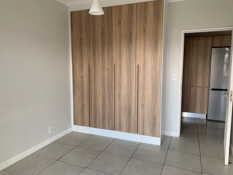 A clean, empty bedroom featuring modern wood-finish built-in cupboards and neutral tiled flooring, with a doorway leading to an adjacent kitchen area.
