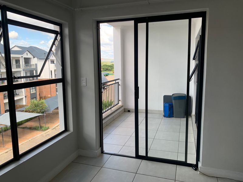 A bright, tiled balcony accessible via a modern black-framed sliding glass door, featuring a metal balustrade and views of the surrounding apartment complex.