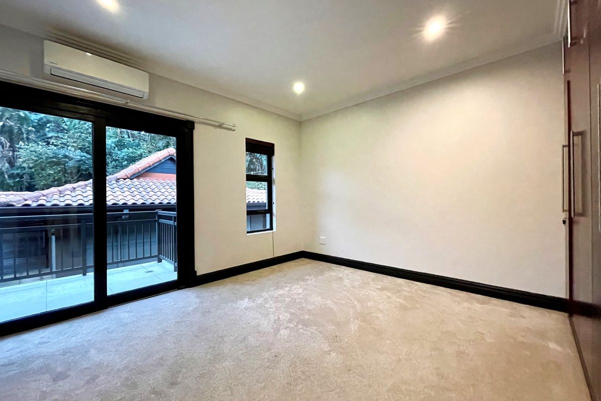 A bright, empty bedroom featuring neutral carpeting, built-in cupboards, recessed lighting, and a sliding glass door leading to a private balcony.