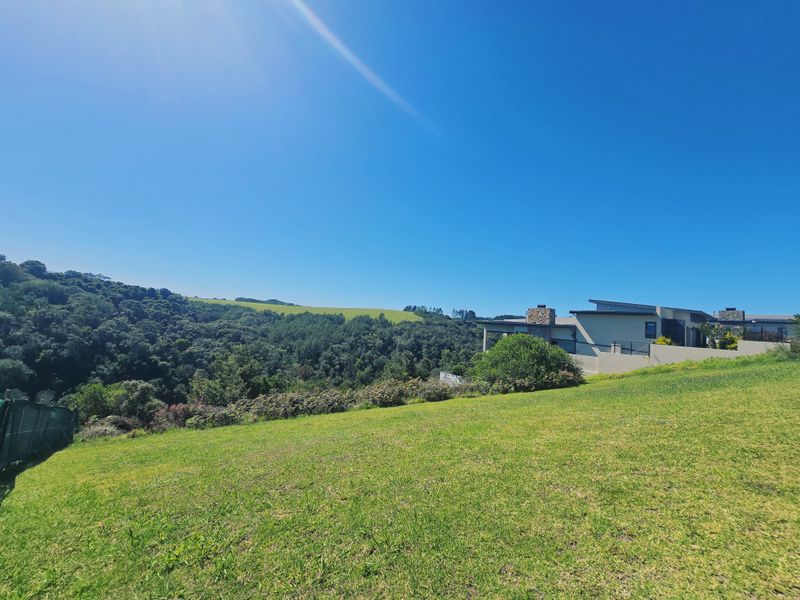 A scenic view of a sloped residential plot featuring lush green grass and surrounding natural landscape with a neighboring modern home visible.