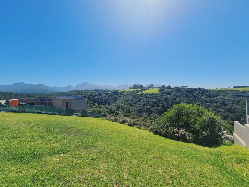A scenic view overlooking a sloped grassy plot with expansive mountain and forest vistas in the background.