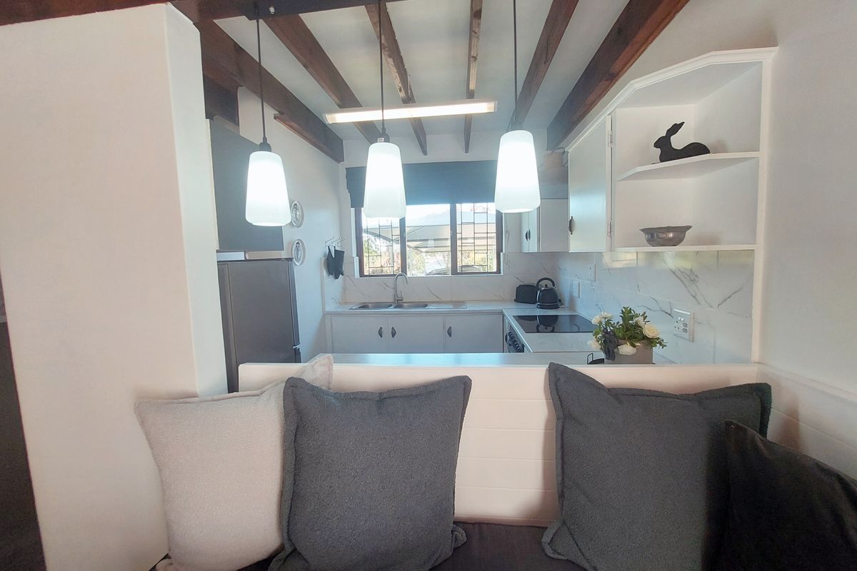 A compact kitchen featuring exposed timber ceiling beams, white cabinetry, marble-look backsplash, and modern pendant lighting, overlooking an adjacent living area.