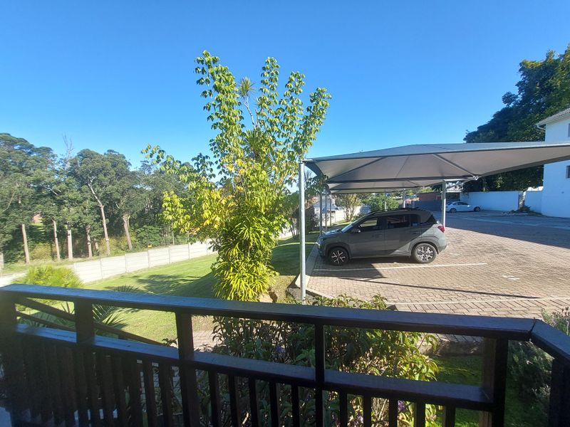 View from a balcony overlooking a paved parking area with shade cloth carports, a lawn, and surrounding trees.