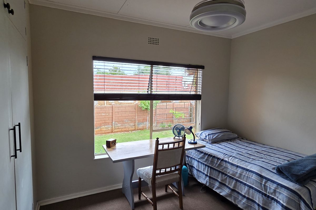 A functional bedroom featuring built-in cupboards, a window with venetian blinds, and a ceiling light fixture, overlooking the rear garden area.