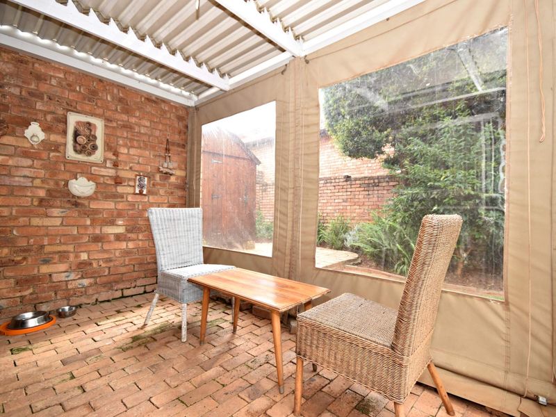 A covered patio area featuring exposed brick walls, brick paver flooring, and protective roll-up canvas blinds overlooking the rear garden.