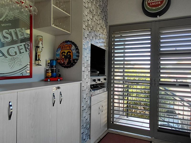 A compact home bar area featuring built-in cabinetry, a decorative tiled splashback, and shutter doors leading to an outdoor space.