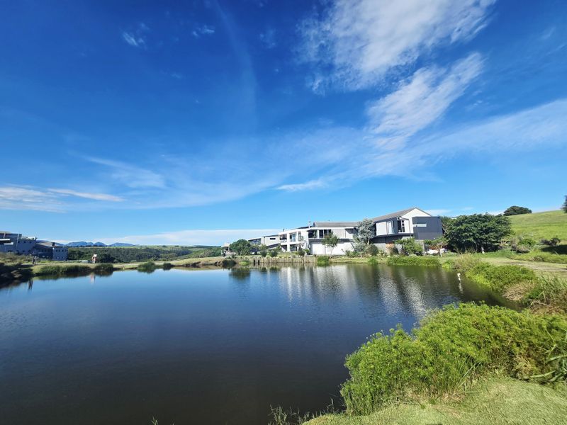 A scenic view of a tranquil pond set against a backdrop of modern residential architecture and rolling green hills.