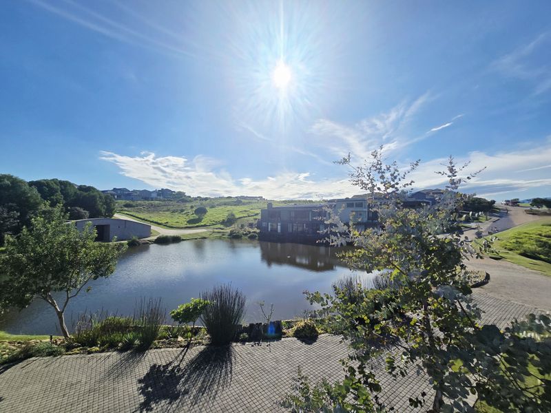 A scenic view overlooking a tranquil pond and landscaped estate grounds, featuring paved pathways and established greenery under a bright sunny sky.