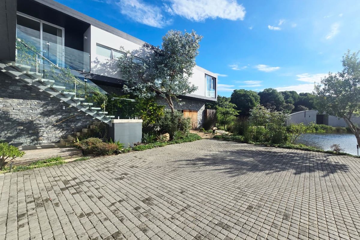 Modern architectural rear exterior featuring stone cladding, a glass balustrade staircase, and a paved driveway overlooking a tranquil pond.