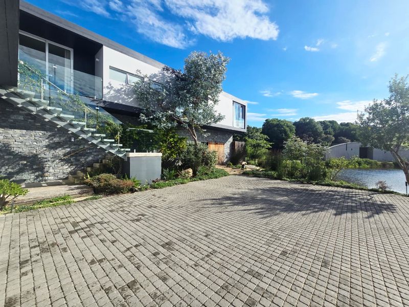 Modern architectural rear exterior featuring stone cladding, a glass balustrade staircase, and a paved driveway overlooking a tranquil pond.