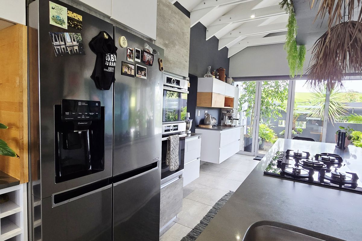 Modern kitchen featuring a large island with gas hob, built-in ovens, sleek cabinetry, and high vaulted ceilings with exposed rafters leading to outdoor access.