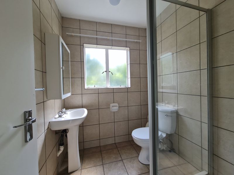 A functional bathroom featuring a pedestal basin, a glass-enclosed shower, and a toilet, finished with neutral wall and floor tiling.