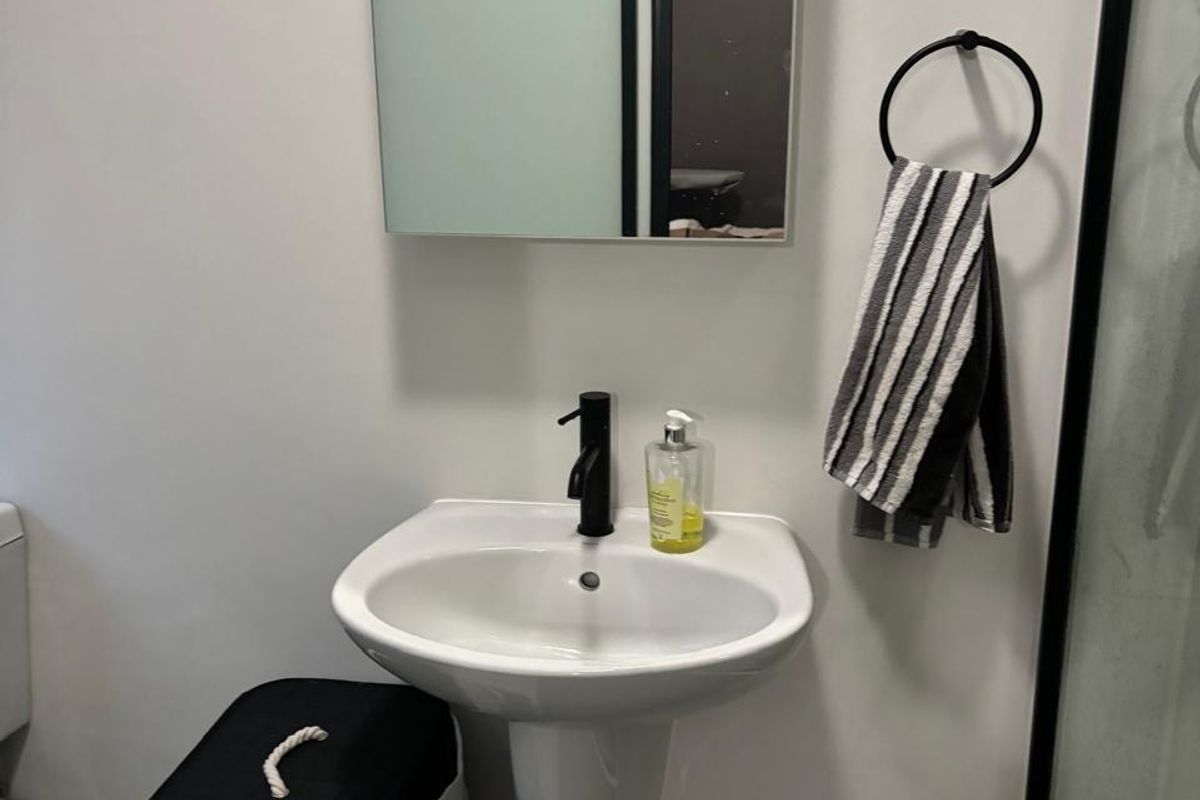 A modern bathroom featuring a white pedestal basin with a black mixer tap, a wall-mounted mirror, and a glass-enclosed shower.