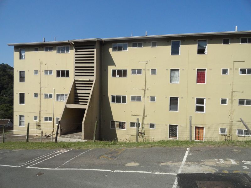 Exterior view of a multi-story apartment block featuring a central stairwell, asphalt parking area, and wire mesh perimeter fencing.