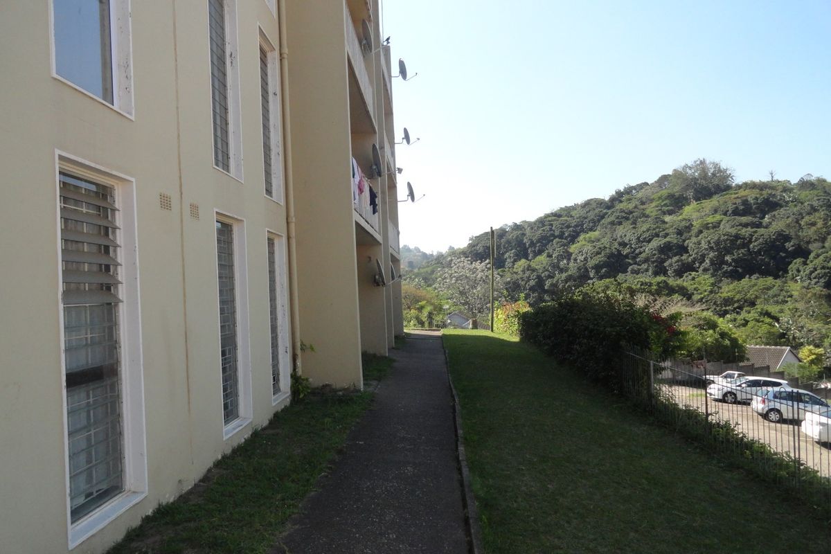Exterior view of the apartment building showing the rear walkway, louvre windows, and surrounding green landscape.