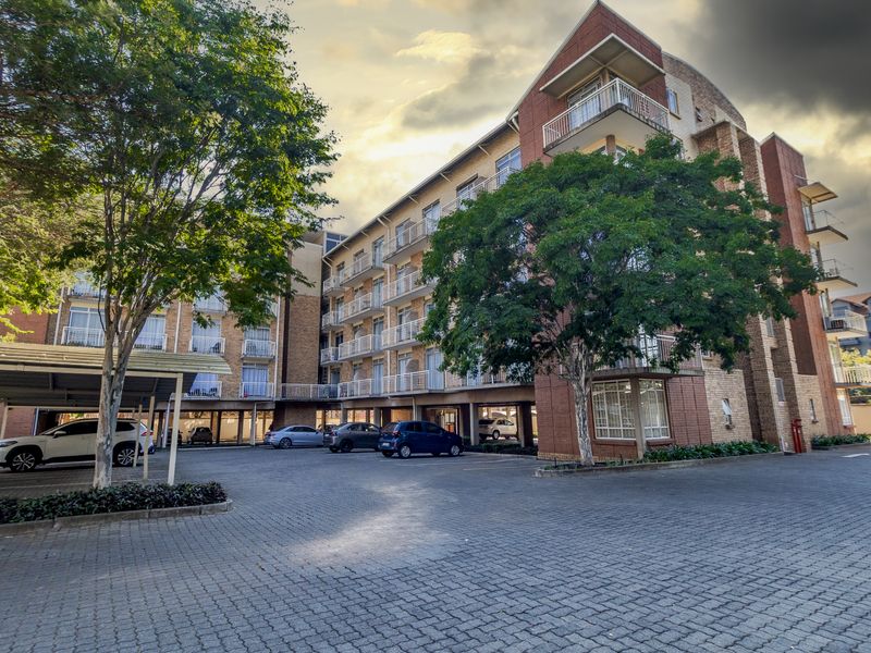 Exterior view of a multi-story apartment complex featuring brick facade, private balconies, and a paved parking area with covered bays.