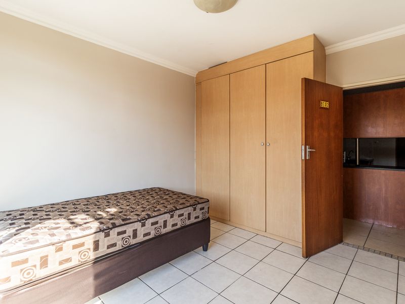 A bedroom featuring tiled flooring, neutral walls, and a built-in wooden wardrobe, with an adjacent view into a kitchen area.