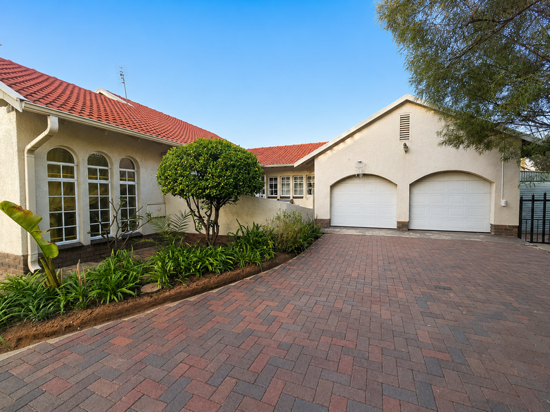 Front elevation of a well-maintained house featuring a double garage, paved driveway, terracotta roof tiles, and arched windows with landscaped garden beds.