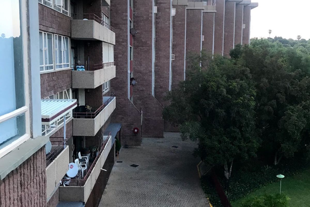 An elevated view of the townhouse complex rear facade, featuring brick construction, multiple balconies, and a paved driveway adjacent to a landscaped garden area.