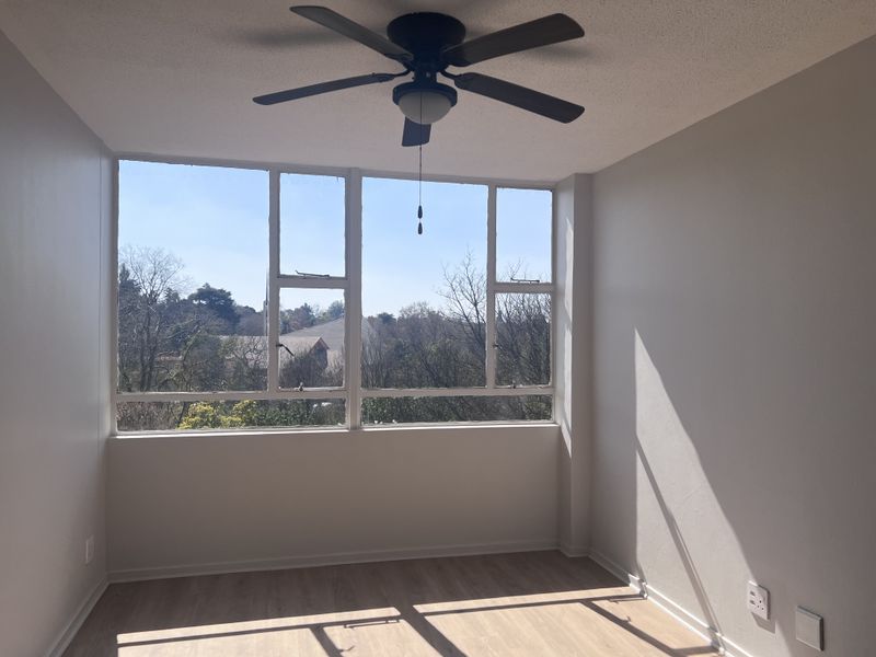 A bright, empty bedroom featuring laminate flooring, a ceiling fan, and large windows offering a pleasant outdoor view.