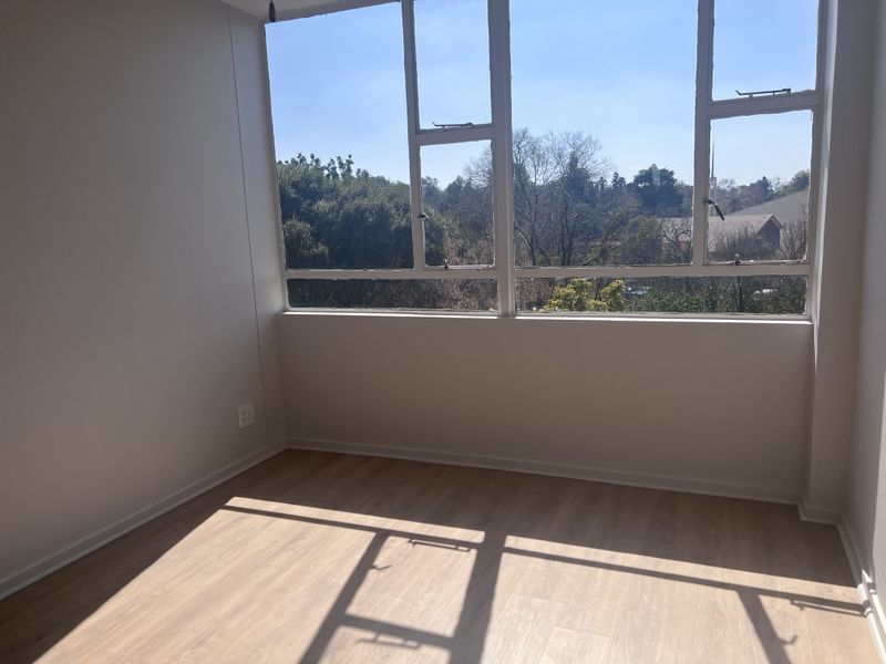 A bright, empty bedroom featuring modern laminate flooring, neutral walls, and a large window offering pleasant views of the surrounding greenery.