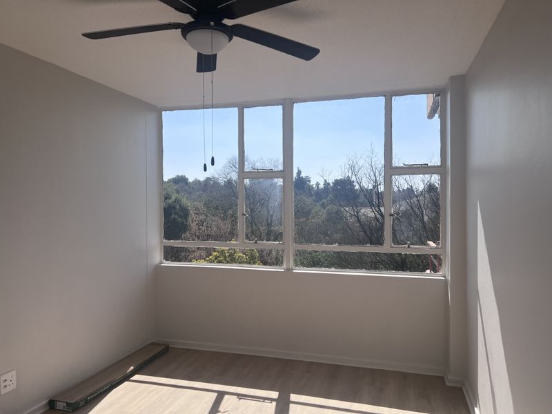 A bright, empty bedroom featuring modern laminate flooring, a ceiling fan, and large windows offering scenic outdoor views.
