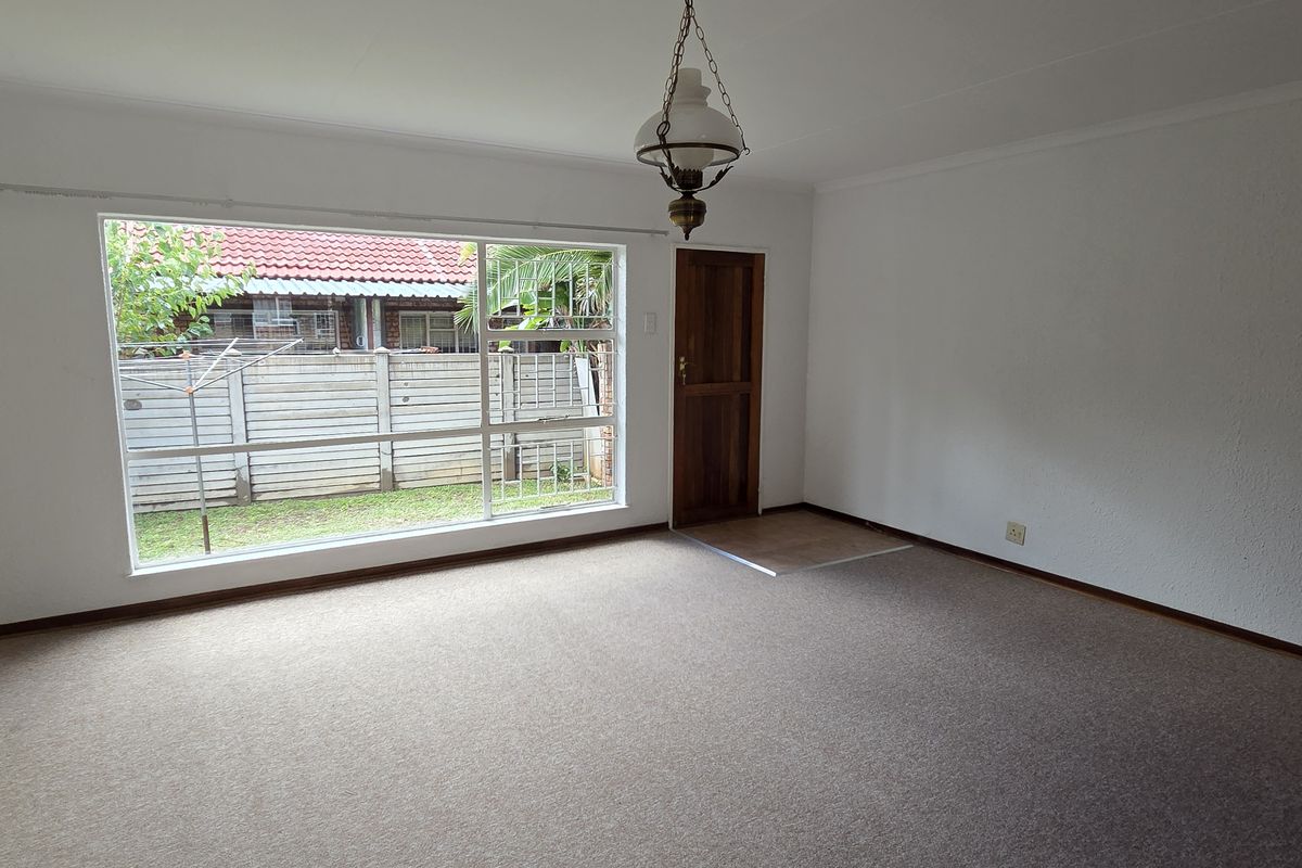 A spacious, empty living room featuring neutral walls, carpet flooring, and a large window providing natural light and a view of the rear garden.