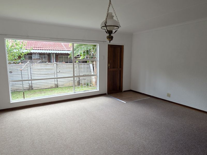 A spacious, empty living room featuring neutral walls, carpet flooring, and a large window providing natural light and a view of the rear garden.