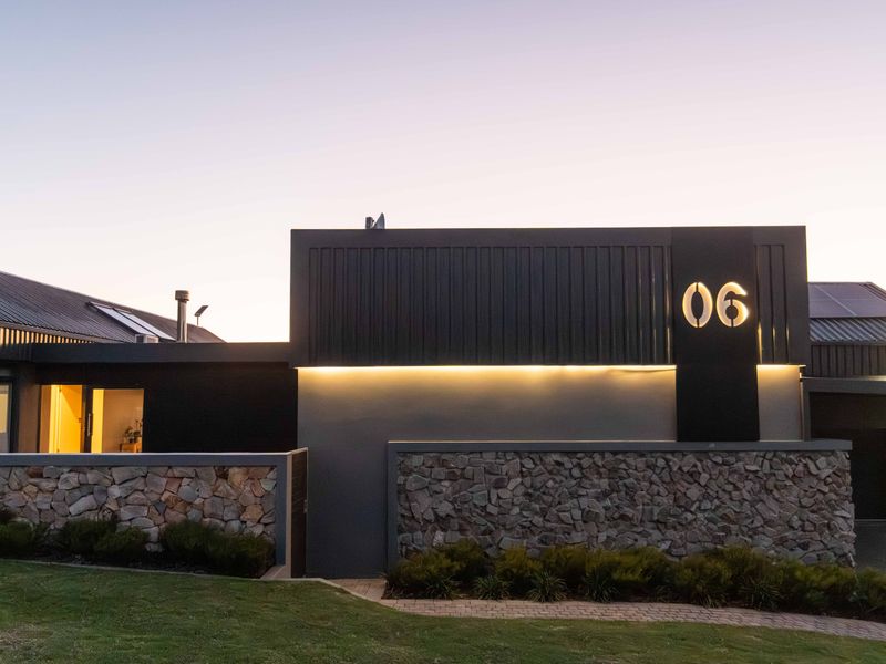 Modern front facade featuring stone cladding, dark metal roofing, integrated solar panels, and illuminated house number signage at dusk.
