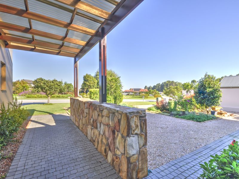 A covered outdoor patio area featuring a stone wall, timber pergola structure, and paved flooring, overlooking a landscaped garden and gravel driveway.