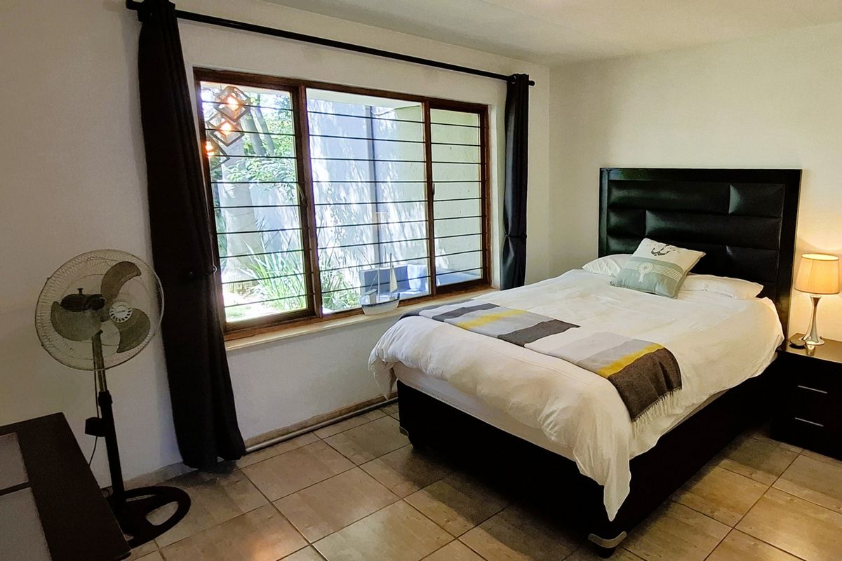 A bedroom featuring tiled flooring and a large window with security bars, providing natural light and a view of the exterior garden area.