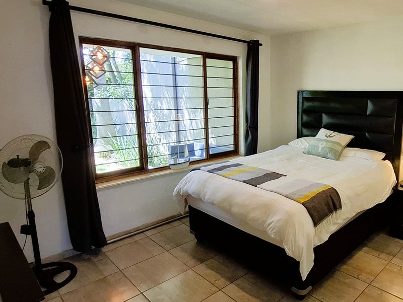 A bedroom featuring tiled flooring and a large window with security bars, providing natural light and a view of the exterior garden area.