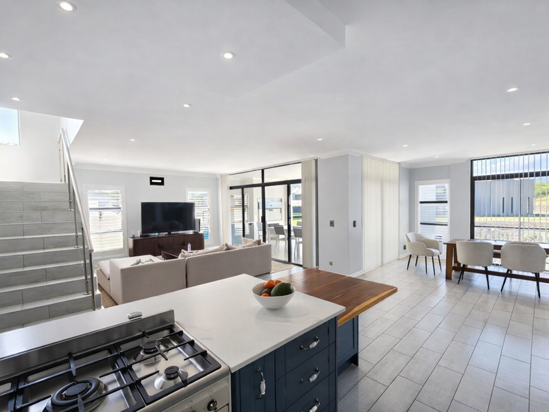 Spacious open-plan living area featuring a modern kitchen with gas cooktop, stone island, tiled floors, and a sleek staircase leading to the upper level.