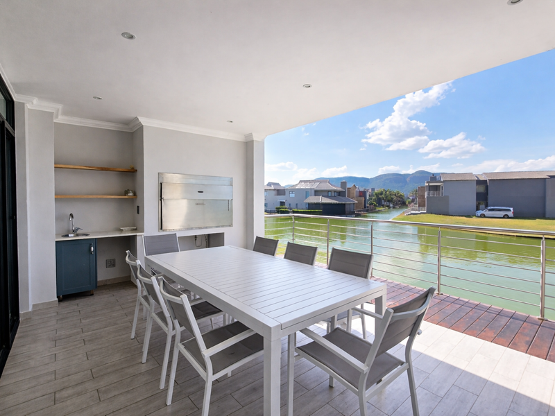 Covered patio featuring a built-in braai, kitchenette with sink, and stainless steel balustrade, offering scenic waterfront and mountain views.