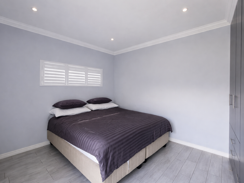 A modern bedroom featuring light grey walls, tiled flooring, built-in cupboards, and plantation shutters, illuminated by recessed downlights.