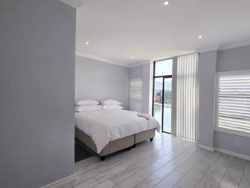 A bright, modern bedroom featuring tiled flooring, recessed lighting, and large glass doors with vertical blinds, offering natural light and views.