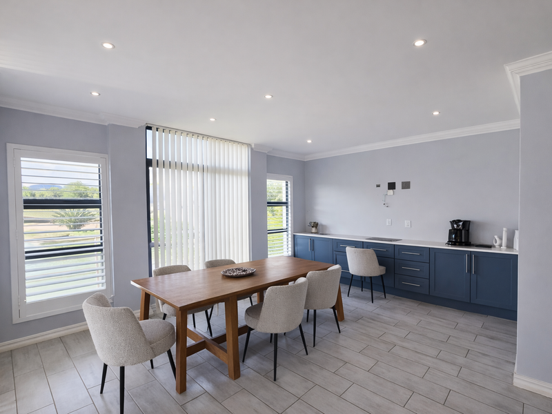 Modern open-plan dining and kitchen area featuring blue cabinetry, stone countertops, tiled flooring, and large windows with vertical blinds.
