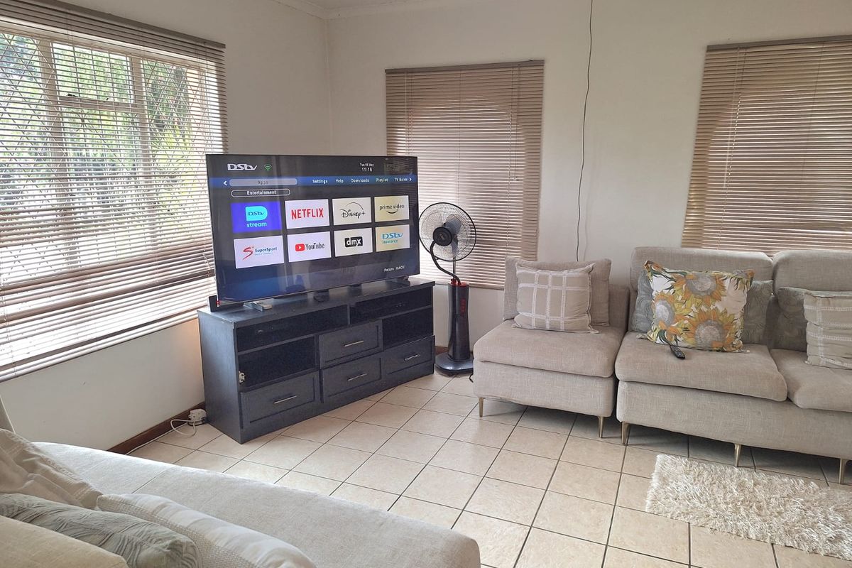 A living room featuring tiled flooring, multiple windows with venetian blinds, and a ceiling fan, providing a functional and well-lit space.