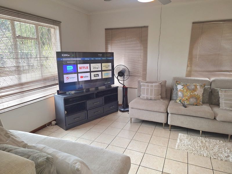 A living room featuring tiled flooring, multiple windows with venetian blinds, and a ceiling fan, providing a functional and well-lit space.