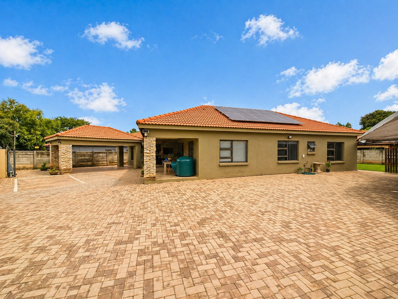 Spacious rear exterior view featuring a large paved driveway, covered patio area, stone-clad pillars, and roof-mounted solar panels.