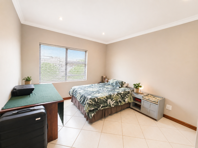A well-lit bedroom featuring neutral wall tones, tiled flooring, and a large window with venetian blinds.