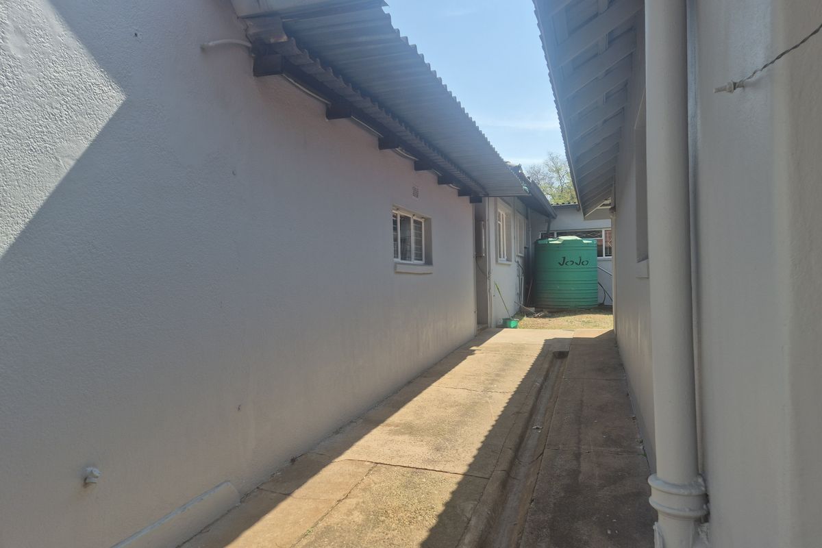 A narrow side passage between building structures featuring a concrete walkway, drainage channel, and a green water storage tank.