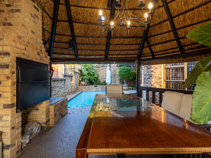 Covered outdoor patio featuring a thatched roof, built-in braai, and tiled flooring, overlooking a private swimming pool area.