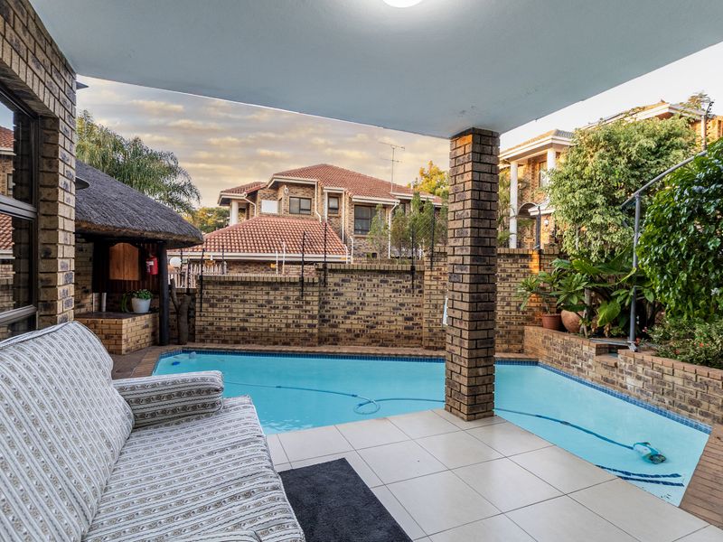 Outdoor entertainment area featuring a covered patio overlooking a swimming pool, brick boundary walls, and a small thatched lapa structure.