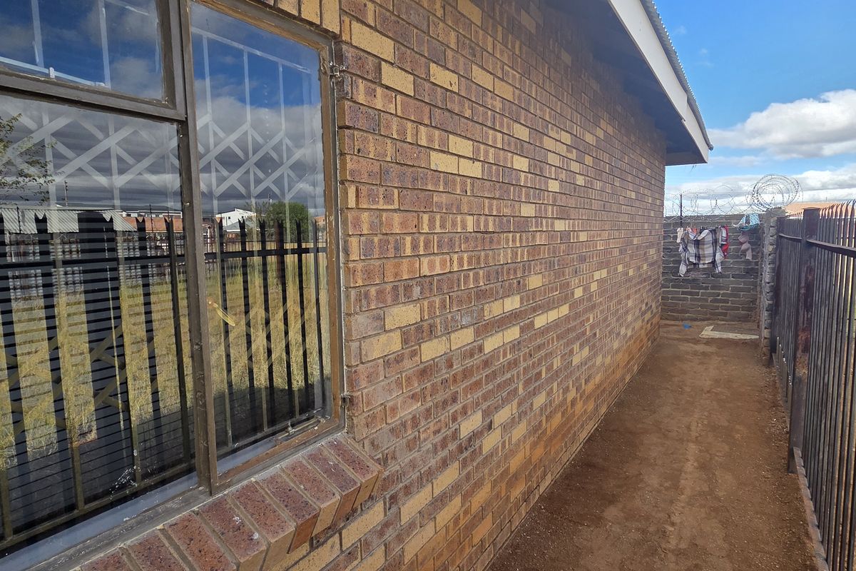 Side exterior view of the house featuring face brick walls, steel security window bars, and metal perimeter fencing with razor wire.