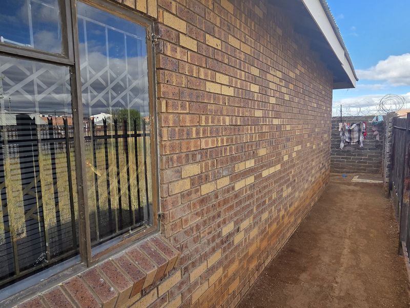 Side exterior view of the house featuring face brick walls, steel security window bars, and metal perimeter fencing with razor wire.